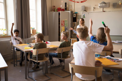 Elever i ett klassrum räcker upp handen och vill svara på lärarens frågor.enom att räcka